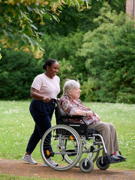 Eine Mitarbeiterin unternimmt mit einer Rollstuhlfahrerin einen Spaziergang im Park. Foto: LWL/Nikolaus Urban